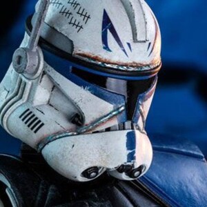 May include: A detailed close-up of a Star Wars Clone Trooper helmet. The helmet is primarily white with blue accents and markings, including tally marks. The image showcases the helmet's design and battle-worn appearance.