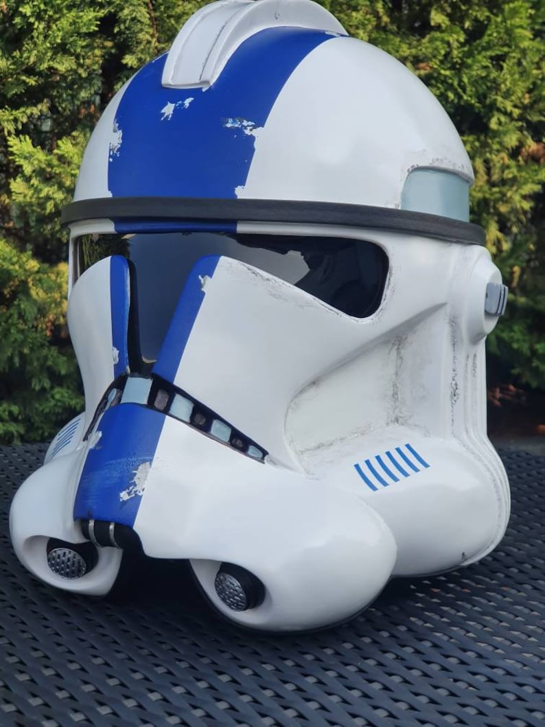 May include: A white and blue helmet with a visor, reminiscent of a clone trooper. The helmet features blue stripes and accents, with a weathered appearance. The design includes details like a chin piece and side attachments.