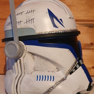 May include: A white and blue helmet with a weathered appearance, featuring blue accents and a stylized design. The helmet has a series of tally marks and a blue emblem. The helmet is a prop or costume piece.