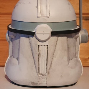 May include: A white and gray helmet with a rounded top, featuring a gray band and black trim. The helmet has a rectangular visor and a cylindrical attachment on the side. The surface shows signs of wear, suggesting a battle-worn aesthetic.