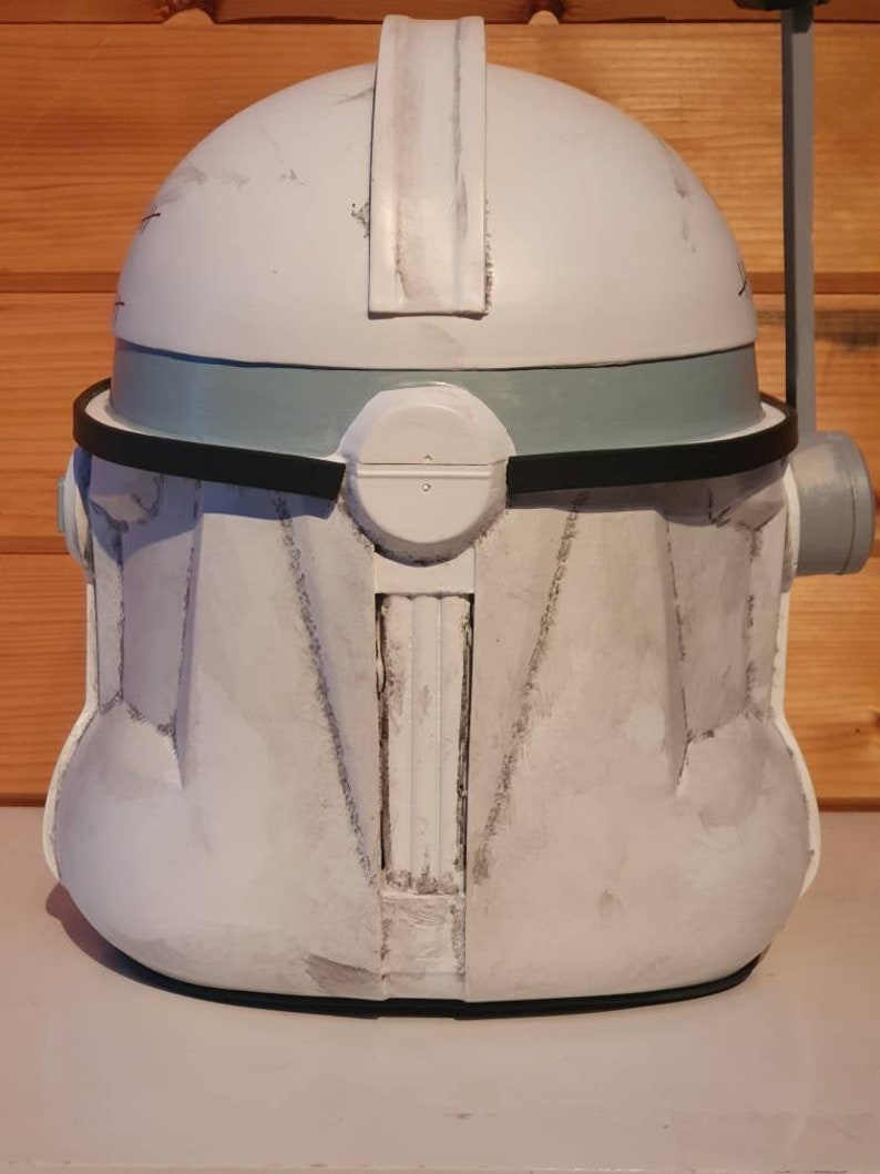 May include: A white and gray helmet with a rounded top, featuring a gray band and black trim. The helmet has a rectangular visor and a cylindrical attachment on the side. The surface shows signs of wear, suggesting a battle-worn aesthetic.