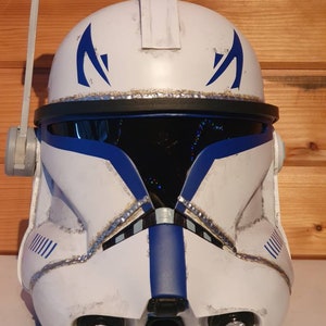 May include: A white and blue helmet with a visor, reminiscent of a clone trooper. The helmet features blue markings and accents, with a silver trim around the visor. It has a weathered appearance, suggesting battle use.