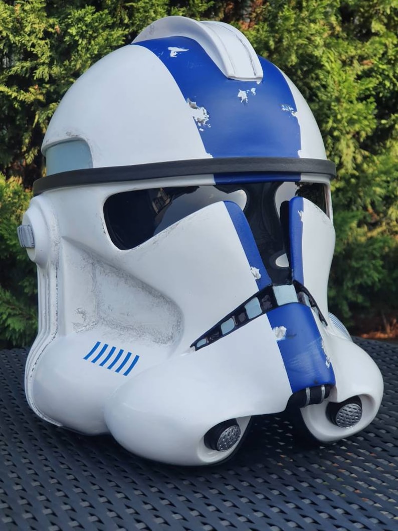 May include: A white and blue helmet with a visor and a rounded top. The helmet has blue stripes and accents, with some areas showing a distressed finish. The helmet is likely a costume piece or collectible.