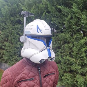 May include: A white and blue helmet with a visor, resembling a clone trooper helmet. The helmet has blue accents and a series of vertical lines on the top. The person is wearing a maroon quilted jacket.