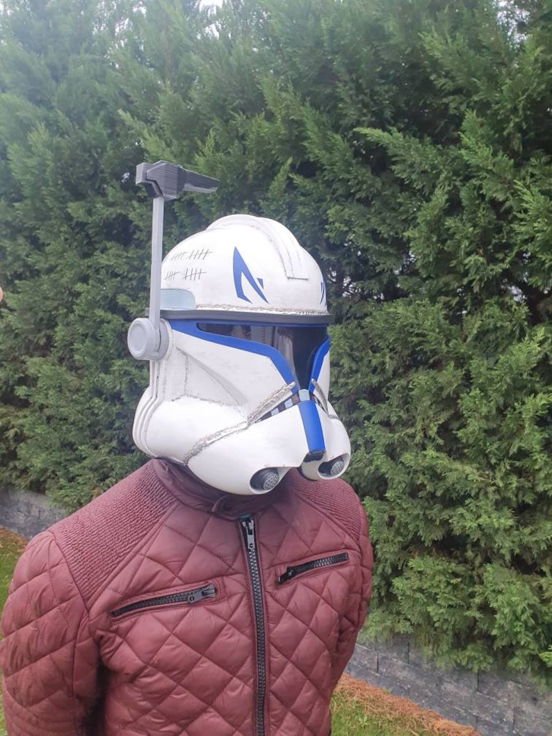 May include: A white and blue helmet with a visor, resembling a clone trooper helmet. The helmet has blue accents and a series of vertical lines on the top. The person is wearing a maroon quilted jacket.