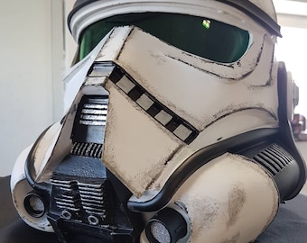 May include: A distressed white and black Stormtrooper helmet with a green visor. The helmet features black detailing and a complex front section. This is a replica of a helmet from the Star Wars films.
