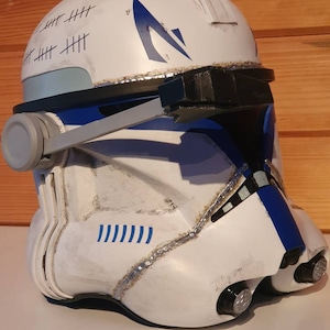 May include: A white and blue helmet with a weathered appearance, featuring blue accents and a stylized emblem. The helmet has a series of tally marks and is designed with a visor and side details, suggesting a futuristic or military theme.