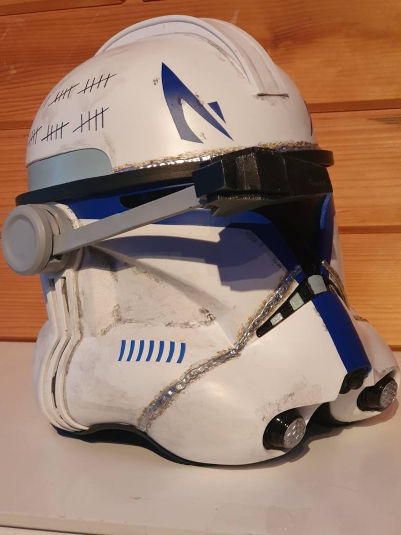 May include: A white and blue helmet with a weathered appearance, featuring blue accents and a stylized emblem. The helmet has a series of tally marks and is designed with a visor and side details, suggesting a futuristic or military theme.