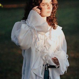 May include: White, ruffled blouse with long, gathered sleeves. The blouse features a high collar and decorative ties. The fabric appears to be a lightweight, textured material. The model is wearing blue jeans.