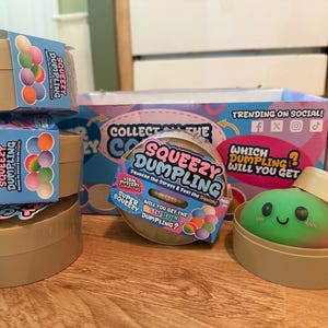 May include: Several Squeezy Dumpling mystery boxes featuring tan containers with blue labels. The labels show colourful dumpling graphics and text including Squeezy Dumpling. One open container reveals a green and pink squishy dumpling toy.