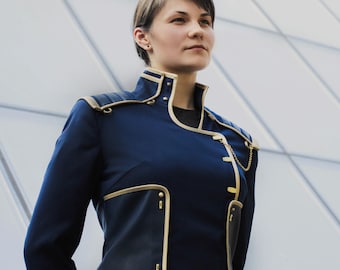 Op de afbeelding: Een donkerblauw jasje in militaire stijl met een hoge kraag, gouden biezen en gestructureerde schouderstukken. Het kledingstuk heeft een sluiting aan de voorzijde met gouden accenten, een klein gouden kettinkje en geribbelde mouwboorden.