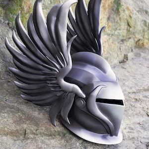 May include: A silver helmet with a winged design. The helmet has a visor and a pointed top. The wings are detailed and have a textured surface.