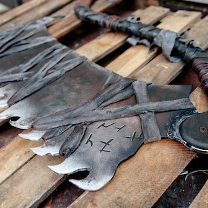 Op de afbeelding: Een groot, gebogen metalen zaagblad met een gekartelde rand, een donkere, verweerde afwerking en omwikkeld met stroken grijze stof. Het blad toont gegraveerde runensymbolen en is bevestigd aan een handvat met donkere leren koordbindingen.