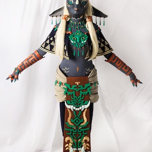 Może przedstawiać: Szczegółowy kostium cosplay z czarną maską z rogami i blond włosami. Strój zawiera granatowy top z geometrycznymi wzorami, pomarańczowe i złote opaski na ramiona oraz brązową i zieloną spódnicę. Kostium ma motyw plemienny.