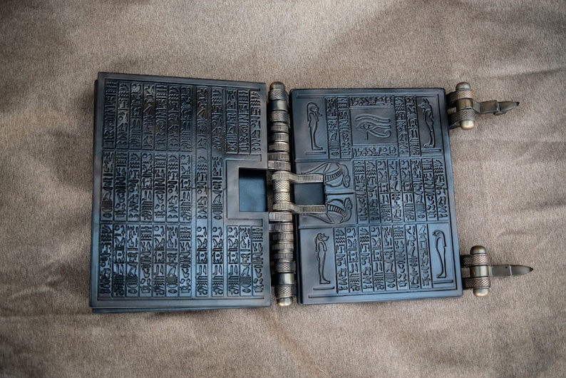 Puede incluir: Un libro de metal negro con intrincados tallados de jerogl&iacute;ficos y un s&iacute;mbolo del ojo de Horus. El libro est&aacute; cerrado y tiene una bisagra de metal.