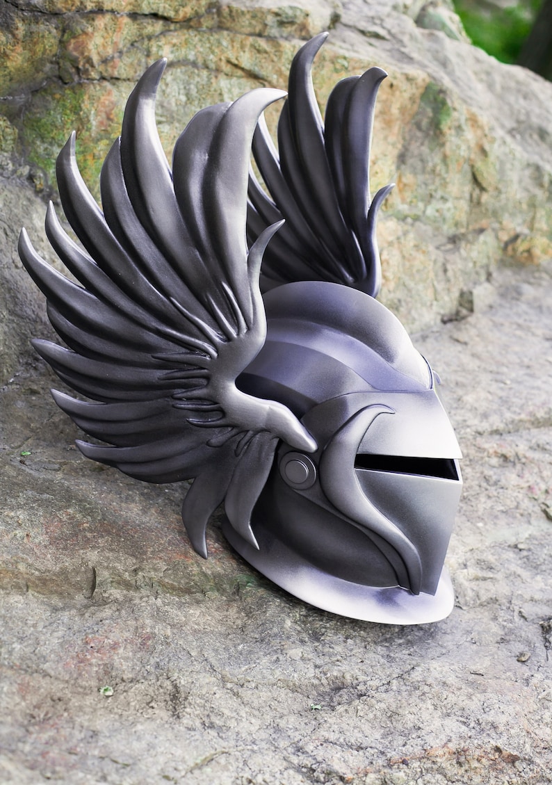 May include: A silver helmet with a winged design. The helmet has a visor and a pointed top. The wings are detailed and have a textured surface.