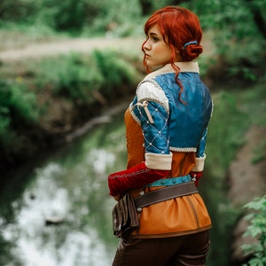 May include: A woman with red hair wearing a blue and brown costume with a leather belt and a brown bag. She is standing in a forest by a stream.