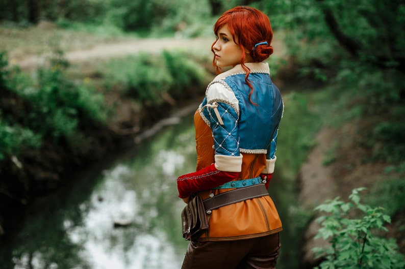 May include: A woman with red hair wearing a blue and brown costume with a leather belt and a brown bag. She is standing in a forest by a stream.
