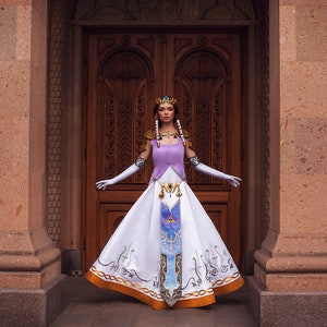 May include: A woman wearing a white and purple dress with gold accents and a crown stands in front of a wooden door. The dress features a design of a triangle with a bird inside, and a pattern of gold lines and swirls.