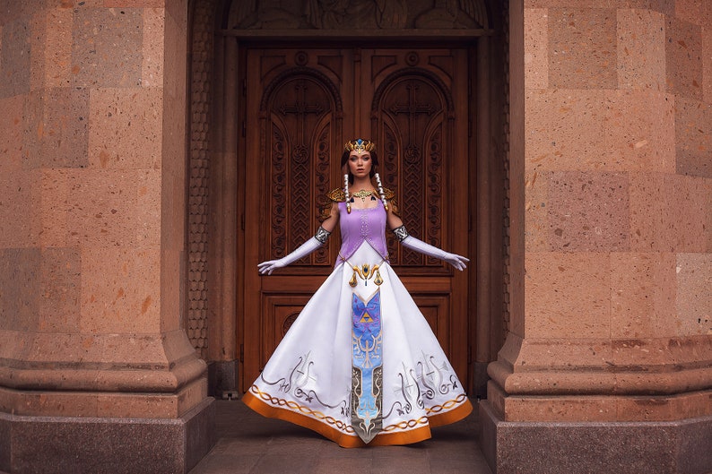 May include: A woman wearing a white and purple dress with gold accents and a crown stands in front of a wooden door. The dress features a design of a triangle with a bird inside, and a pattern of gold lines and swirls.
