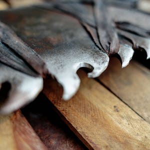 Op de afbeelding: Een grote, verweerde metalen boomzaag met opvallende, scherpe, gekartelde tanden. Het blad vertoont sporen van oxidatie en roest, met donkere, versleten stoffen stroken om delen van het metaal gewikkeld, rustend tegen een stapel ruwe houten planken.