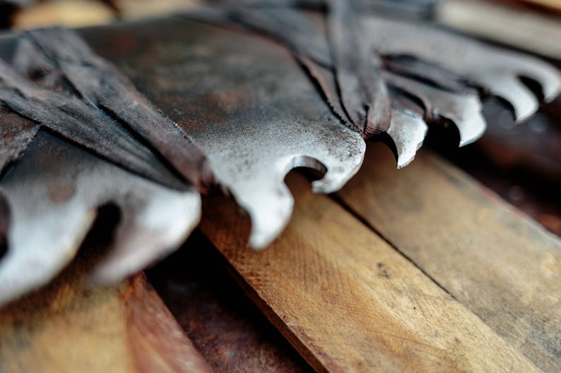 Op de afbeelding: Een grote, verweerde metalen boomzaag met opvallende, scherpe, gekartelde tanden. Het blad vertoont sporen van oxidatie en roest, met donkere, versleten stoffen stroken om delen van het metaal gewikkeld, rustend tegen een stapel ruwe houten planken.