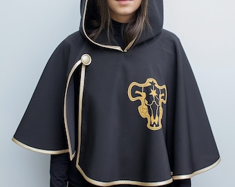 Op de afbeelding: Zwarte cape met capuchon met gouden rand en een gouden embleem. De cape heeft een knoopsluiting en een capuchon. Het embleem is een gestileerde stier. De cape wordt gedragen over een zwart shirt met lange mouwen en een zwarte broek.