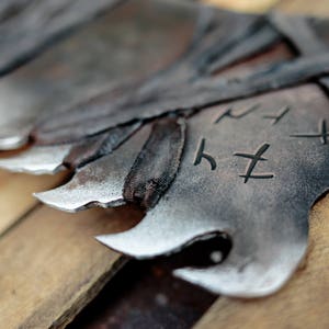 Op de afbeelding: Een reeks donkere, gesmede metalen bladen met gekartelde, puntige randen en een verweerde, getextureerde afwerking. Elk blad is voorzien van donker, omwikkeld materiaal bij de basis en duidelijke, gegraveerde runensymbolen op het metaal.
