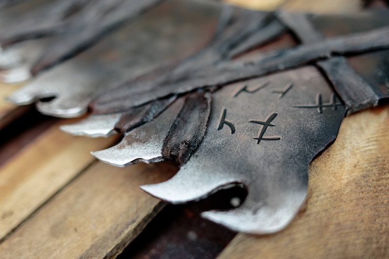 Op de afbeelding: Een reeks donkere, gesmede metalen bladen met gekartelde, puntige randen en een verweerde, getextureerde afwerking. Elk blad is voorzien van donker, omwikkeld materiaal bij de basis en duidelijke, gegraveerde runensymbolen op het metaal.