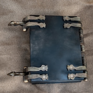 Puede incluir: Un diario de cuero negro encuadernado con cierres met&aacute;licos ornamentados y una columna vertebral decorativa. El diario tiene una est&eacute;tica vintage y es perfecto para escribir o llevar un diario.