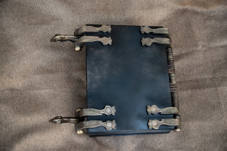 Puede incluir: Un diario de cuero negro encuadernado con cierres met&aacute;licos ornamentados y una columna vertebral decorativa. El diario tiene una est&eacute;tica vintage y es perfecto para escribir o llevar un diario.