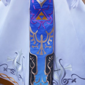 May include: A long, blue and gold fabric sash with a stylised design of a bird and a triangle. The sash is attached to a white dress with gold trim.