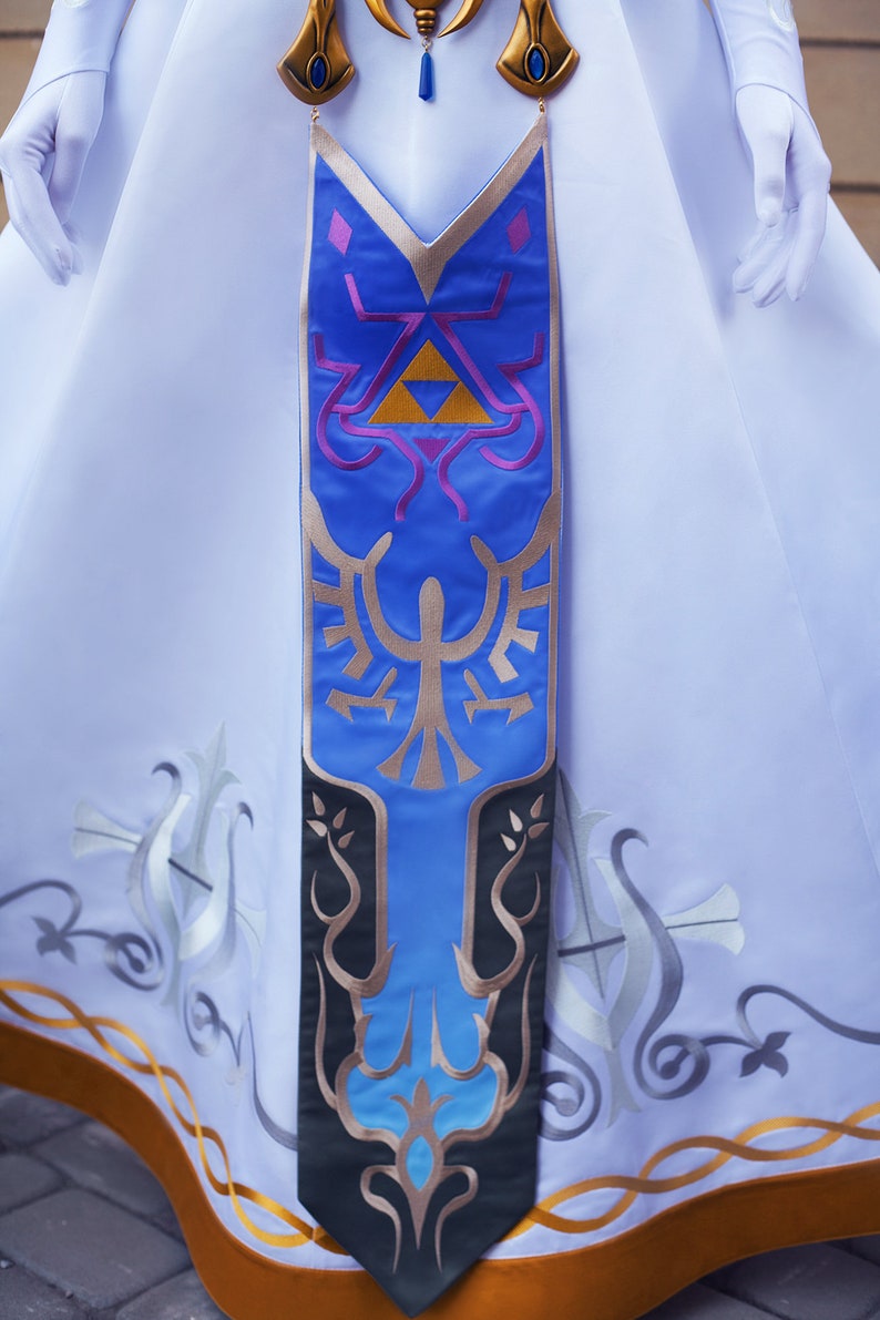 May include: A long, blue and gold fabric sash with a stylised design of a bird and a triangle. The sash is attached to a white dress with gold trim.