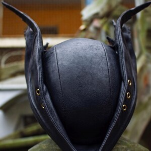 May include: A black leather hat with a unique design. The hat features a rounded crown and extended flaps with brass eyelets. The hat is sitting on a stone surface, with a blurred background.