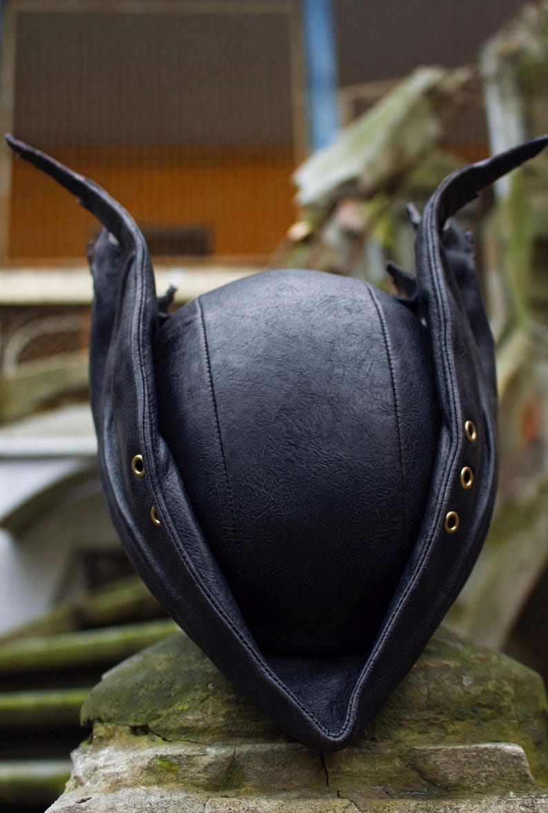 May include: A black leather hat with a unique design. The hat features a rounded crown and extended flaps with brass eyelets. The hat is sitting on a stone surface, with a blurred background.