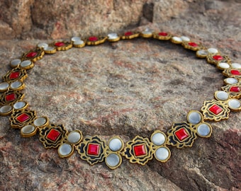 May include: A gold necklace with red and white stones. The necklace is laid out in a circle on a brown, textured surface.