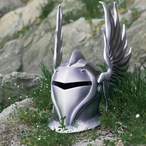 May include: A silver knight helmet with a winged design. The helmet is sitting on a bed of green grass and rocks.