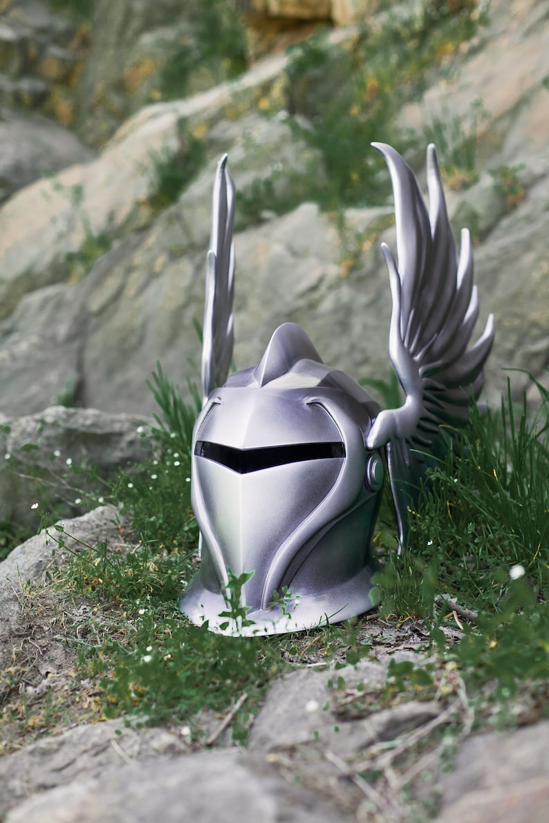 May include: A silver knight helmet with a winged design. The helmet is sitting on a bed of green grass and rocks.