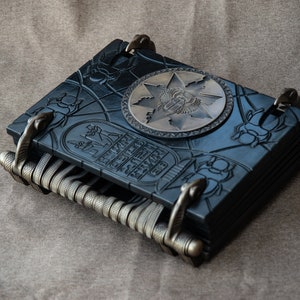 Puede incluir: Un diario encuadernado en cuero negro con tallas intrincadas y un cierre de metal. La portada presenta un dise&ntilde;o en relieve de un escarabajo egipcio y jerogl&iacute;ficos. Una placa de metal circular con un s&iacute;mbolo egipcio est&aacute; centrada en la portada.