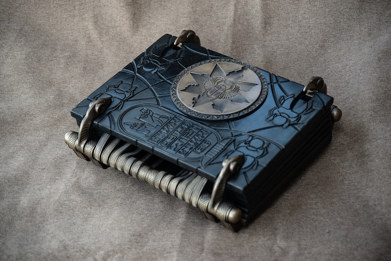 Puede incluir: Un diario encuadernado en cuero negro con tallas intrincadas y un cierre de metal. La portada presenta un dise&ntilde;o en relieve de un escarabajo egipcio y jerogl&iacute;ficos. Una placa de metal circular con un s&iacute;mbolo egipcio est&aacute; centrada en la portada.