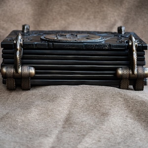 Puede incluir: Un libro encuadernado en cuero negro con cierres de metal adornados y un dise&ntilde;o decorativo en la portada.