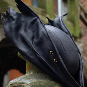 May include: Black leather tricorn hat with a distinctive, pointed brim and two brass eyelets. The hat's design features a rounded crown and a wide, upturned brim, creating a dramatic silhouette. The hat is likely for cosplay or costume use.