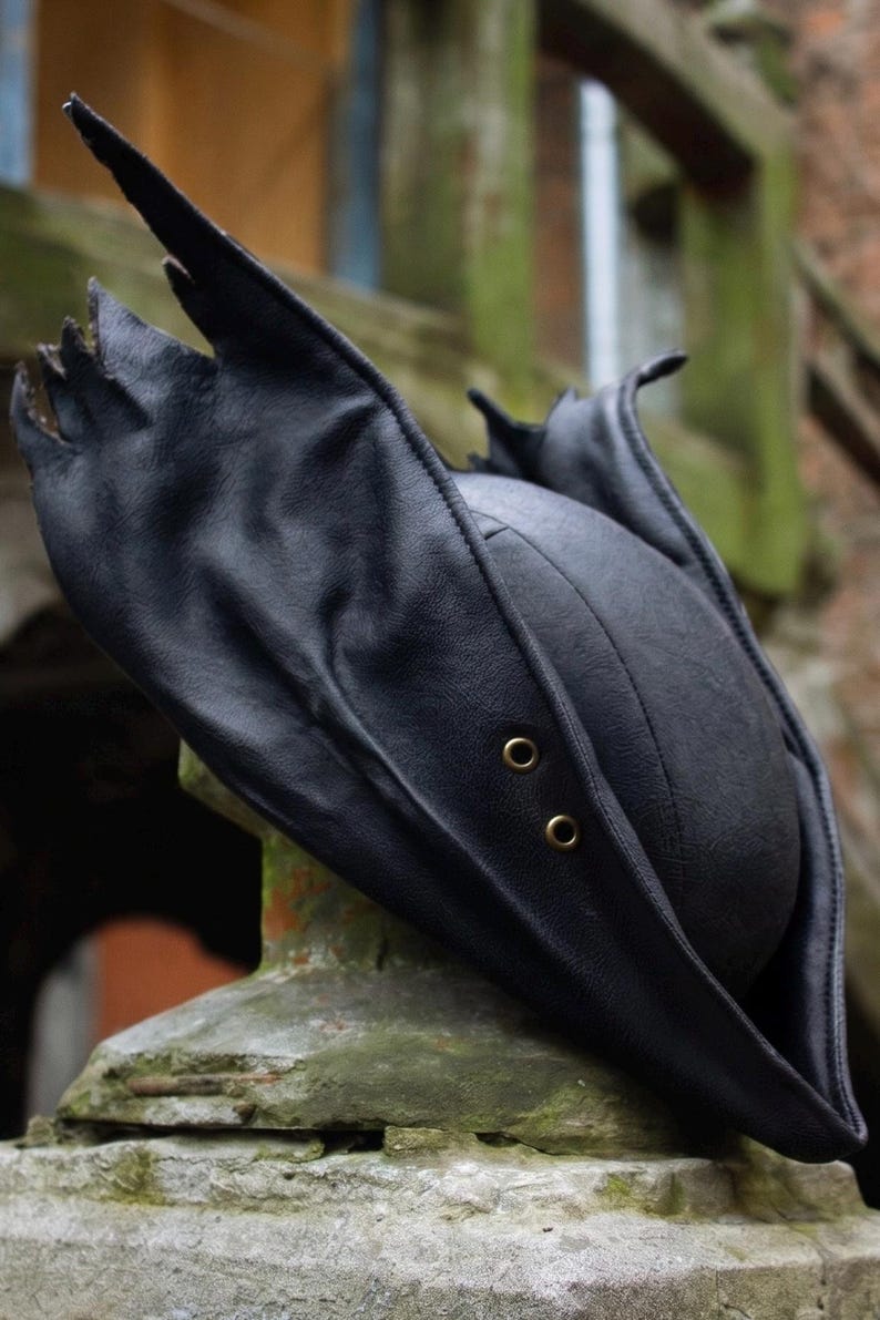 May include: Black leather tricorn hat with a distinctive, pointed brim and two brass eyelets. The hat's design features a rounded crown and a wide, upturned brim, creating a dramatic silhouette. The hat is likely for cosplay or costume use.