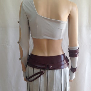 May include: A light gray one-shoulder crop top with a long sleeve featuring cutouts and metal fasteners, paired with a matching pleated skirt. A wide, dark burgundy belt with a hanging strap and matching arm cuffs complete the ensemble.