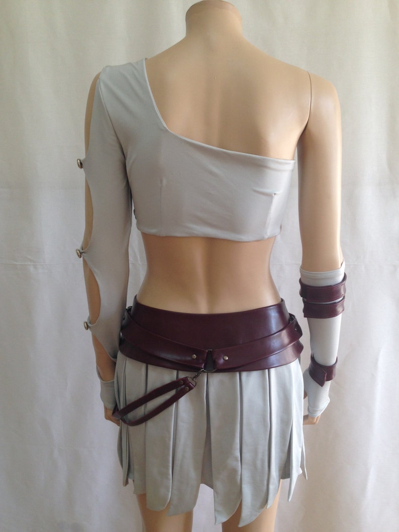 May include: A light gray one-shoulder crop top with a long sleeve featuring cutouts and metal fasteners, paired with a matching pleated skirt. A wide, dark burgundy belt with a hanging strap and matching arm cuffs complete the ensemble.