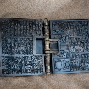Puede incluir: Un libro de metal negro con un dise&ntilde;o de bisagras. El libro est&aacute; cubierto de intrincados tallados de jerogl&iacute;ficos y escarabajos. El libro est&aacute; abierto para revelar m&aacute;s tallados en el interior.