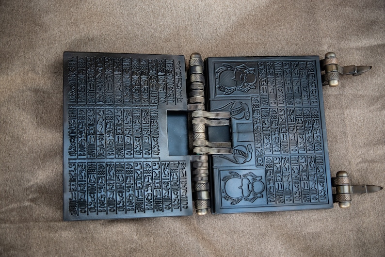 Puede incluir: Un libro de metal negro con un dise&ntilde;o de bisagras. El libro est&aacute; cubierto de intrincados tallados de jerogl&iacute;ficos y escarabajos. El libro est&aacute; abierto para revelar m&aacute;s tallados en el interior.