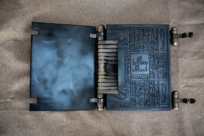 Puede incluir: Un libro negro abierto con una bisagra de metal y tallas intrincadas en la portada. Las tallas incluyen jerogl&iacute;ficos e im&aacute;genes de dioses egipcios.