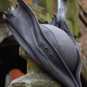 May include: A dark gray, stylized hat with a unique, pointed brim and decorative edging. The hat features a rounded crown and two eyelets on the front. The hat is made of a soft, flexible material.