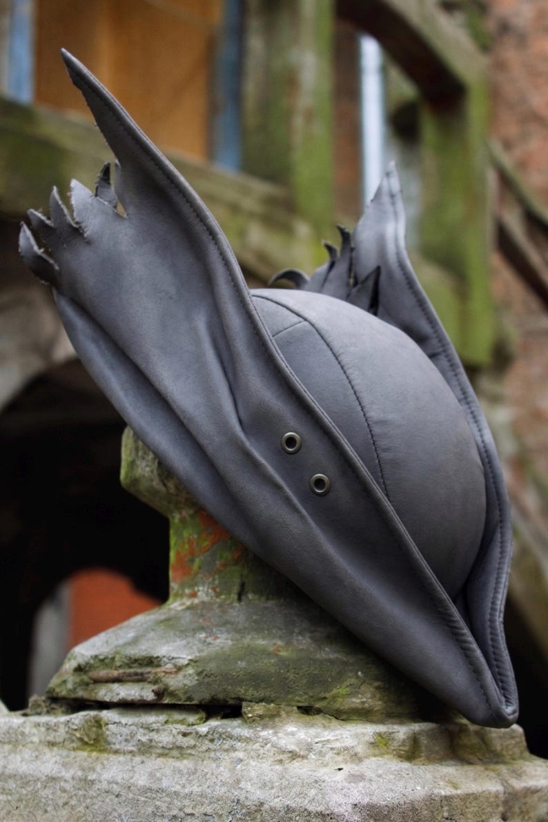 May include: A dark gray, stylized hat with a unique, pointed brim and decorative edging. The hat features a rounded crown and two eyelets on the front. The hat is made of a soft, flexible material.